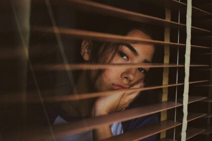 girl thru blinds a dark photo of a girl staring at the camera through wooden blinds. featured on a post about imposter syndrome being sexist and how to overcome imposter syndrome